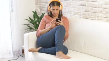Evdeki beyaz koltukta dans eden ve kulaklıkla müzik dinleyen güzel bir genç kadın.