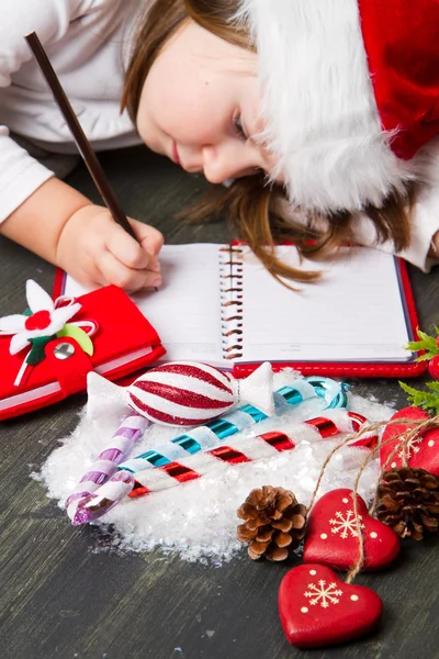 Funny girl in Santa hat writes letter to Santa near christmas decoration — Stock Photo, Image