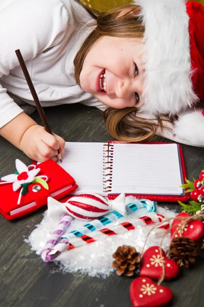 Funny girl in Santa hat writes letter to Santa near christmas decoration — Stock Photo, Image