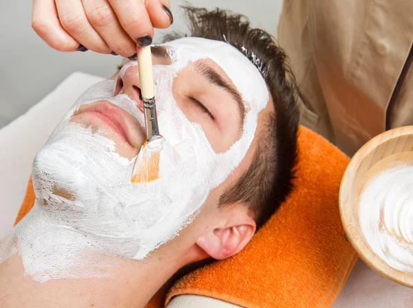 Terapeuta aplicando una mascarilla facial a un hermoso joven en un spa — Foto de Stock