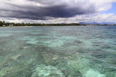 Canlı turkuaz okyanus dalgalanan bulutların dramatik gökyüzünün altında uzak bir tropikal kıyıya uzanıyor.