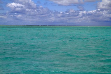 Berrak turkuaz su, kayalıklı beyaz kum kıyısı ve uzak teknelerin yer aldığı huzurlu bir sahil sahnesi.