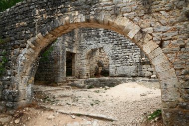 Yıpranmış taş kemerler ve çökmüş ortaçağ duvarları sessiz, güneşli bir avlu oluşturarak kırsal alanda tarihi, çürümeyi ve mimari işçiliği anımsatıyor..