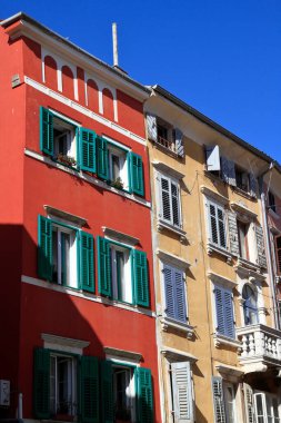 Yıpranmış taş binalar tarafından çerçevelenmiş dar bir Akdeniz geçidi, yeşil kepenkleri ve kır cazibesini, tarihi ve ılık yaz ışığını çağrıştıran çatlak kaldırım taşı yolu olan turuncu bir perdenin yer aldığı.