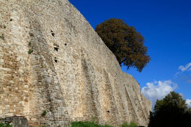 Tarihi bir tahkimattan oluşan yıpranmış bir ortaçağ taş duvarı, temelinde yeşil çimenler ve üzerinde açık mavi bir gökyüzü bulunan görkemli bir şekilde ayakta duruyor..