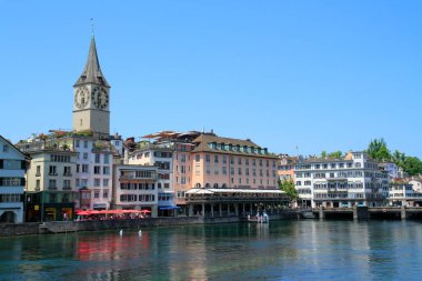 Zürih Limmat Nehri 'nin geniş bir manzarası, tarihi binalar ve parlak mavi gökyüzünün altında kilise kulesiyle dolu canlı bir gezinti alanı..