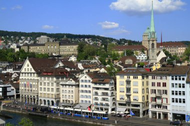 Zürih Limmat Nehri 'nin geniş bir manzarası, tarihi binalar ve parlak mavi gökyüzünün altında kilise kulesiyle dolu canlı bir gezinti alanı..