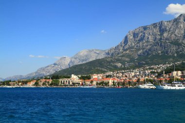 Kırmızı çatı evleri ve marinası olan sakin bir sahil köyü engebeli bir dağ zeminine, ön planda derin mavi bir denize ve berrak bir gökyüzünün altında sıcak bir güneş ışığına sahip sakin ve kaçmayı başarmış..