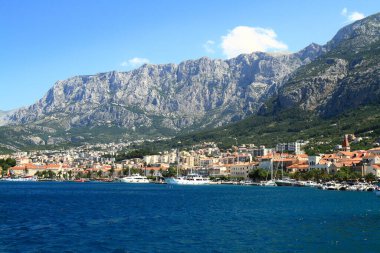 Kırmızı çatı evleri ve marinası olan sakin bir sahil köyü engebeli bir dağ zeminine, ön planda derin mavi bir denize ve berrak bir gökyüzünün altında sıcak bir güneş ışığına sahip sakin ve kaçmayı başarmış..