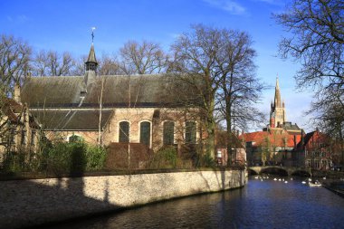 Bruges 'deki güneş ışığı kanalı sahnesinde kırmızı çatıların üzerinde yükselen uzun bir ortaçağ kilise kulesi, yayaların ve yapraksız ağaçların olduğu taş bir köprü yer alıyor.
