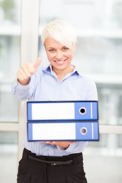 Businesswoman making positive thumb gesture while holding pile o — Stock Photo, Image