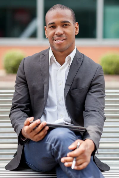 Hombre de negocios africano guapo está sentado en un banco con tableta — Foto de Stock