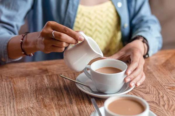 Закрыть руки женщины наливая молоко или сливки в чашку кофе на — стоковое фото