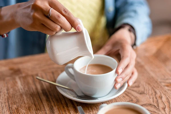 Закрыть руки женщины наливая молоко или сливки в чашку кофе на — стоковое фото