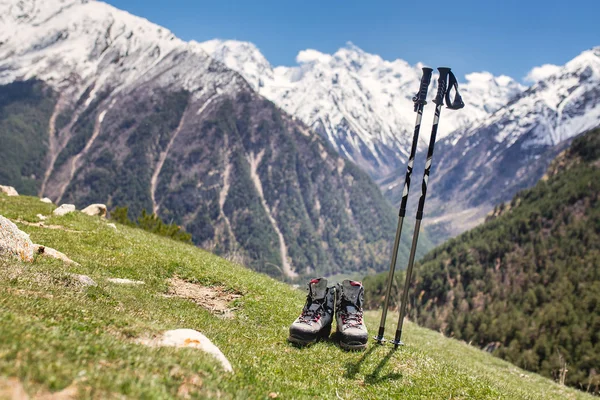 Hiking boots with trekking poles on the grass on the mountain snowy Caucasus range background — Stock Photo, Image