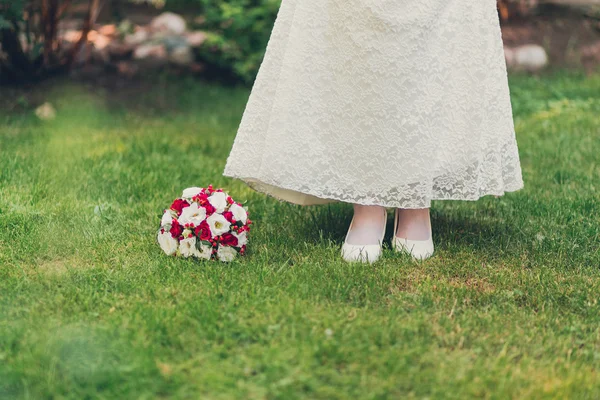 Meisje in witte jurk en witte schoenen staande op groen gras naast een boeket van rode bloemen en groen — Stockfoto