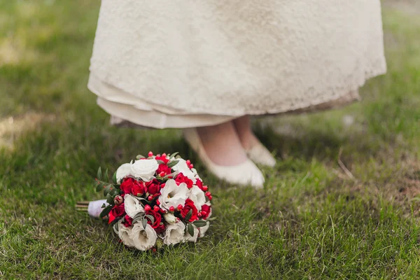 Niña en vestido blanco y zapatos blancos de pie sobre hierba verde junto a un ramo de flores rojas y vegetación — Foto de Stock