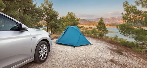 The blue tent is set up on a flat area in the forest near a car. Road trips and camping in the woods