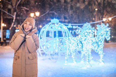 Neşeli bir kadın, Noel ve Yeni Yıl arifesinde çelenkler ve ışıklarla muhteşem bir kış kasabasında mutlu ve eğleniyor. Arka planda atlar olan parlak bir araba.