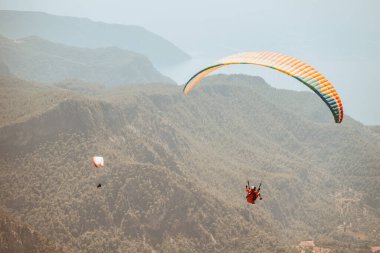 Eğitmen ve öğrenciyle birlikte bir paraglider ormanlık dağ ve tepelerin arka planında süzülüyor.