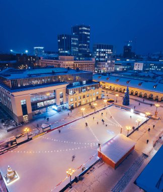 Yeni yıl için dekore edilmiş bir Noel ağacının arka planındaki buz pateni sahasında alışveriş merkezinin yakınlarındaki muhteşem kış şehrinde eğlenen ve kayan insanların hava manzarası.