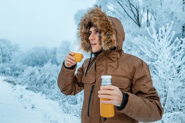 Kapüşonlu neşeli bir adam soğuk kar fırtınası ve donmadan sonra termostan sıcak çay içer.