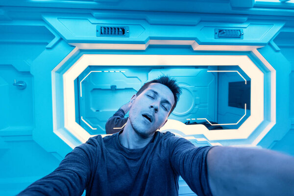 Happy caucasian client of a futuristic capsule hotel takes a selfie near the mirror before going to sleep. The modern interior references to a cryogenic shuttle in a spaceship