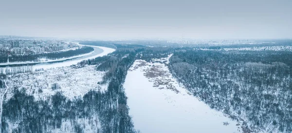 Highrise buildings form a neighboring residential area near a picturesque snow covered river in the city. Drone Aerial view in winter