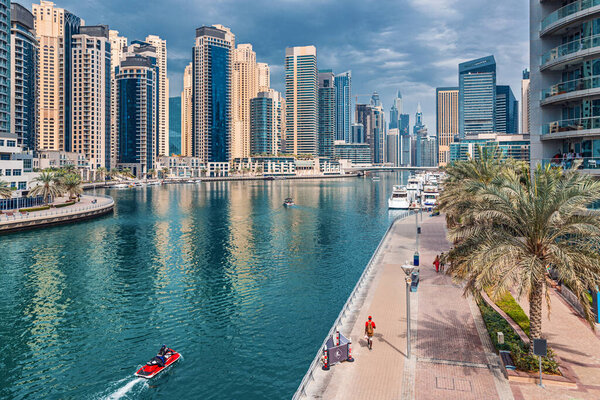 Modern and developed Dubai Marina area with high skyscrapers and commercial and residential real estate. A jet ski is floating on the water and people are walking along the embankment