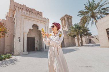 Renkli türbanlı doğulu bir kadın arkadaşlarıyla mesajlaşır ve akıllı bir telefonla eski bir Dubai şehrinin arka planına karşı iletişim kurar.
