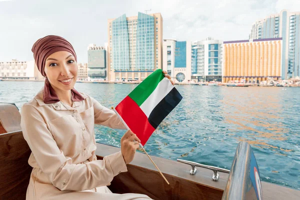 Happy woman with the flag of the United Arab Emirates sits in a boat during a cruise on the Dubai Creek Canal with views of numerous skyscrapers and attractions