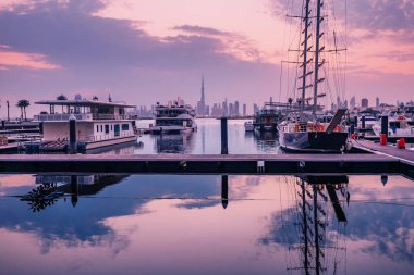 Dubai Creek Marina Limanı 'nda, ünlü Burj Halife' nin silueti ile şehir merkezine bakan yat ve gemiler renkli bir günbatımında park ediliyor.