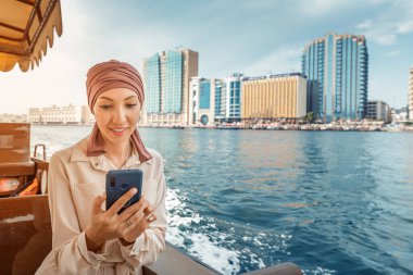 Dubai Creek 'te geleneksel bir Abra Dhow teknesinde dolaşırken akıllı telefonunda uygulama kullanan bordo sarıklı mutlu bir Asyalı kadın..