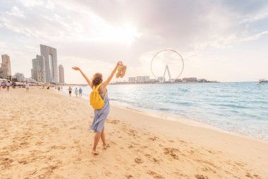 Mutlu kadın turist Dubai 'nin JBR bölgesinde kumlu bir sahilde yalınayak yürüyor ve Ain Ferris Wheel' in panoramik manzarasına hayran.