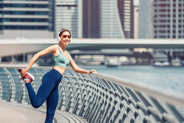 Happy woman wearing headphones stretches the quadriceps muscles of the thigh and warms up before intense running workout on the embankment in Dubai Marina