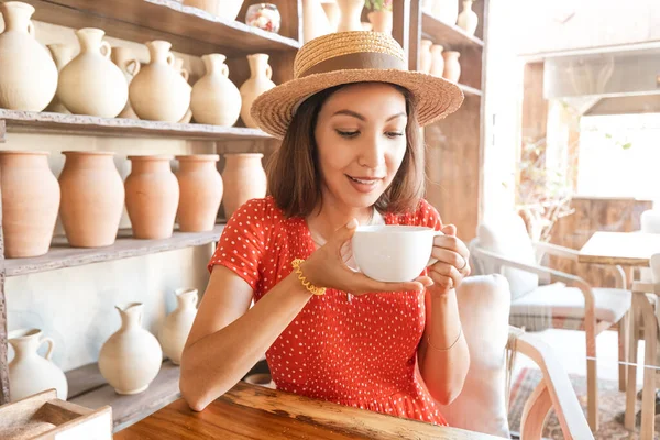 A woman of Indian or Indonesian appearance drinks tea or coffee from a large mug in a cafe against the background of pottery as a decor