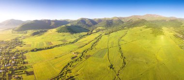 Yeşiller ve köy evlerinin panoramik hava manzarası. Arka planda yükselen güneşin aydınlattığı yüksek dağlar.