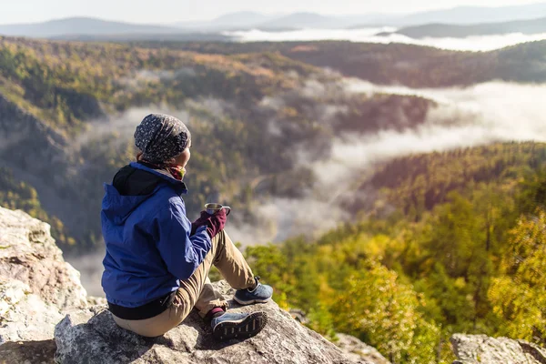 Jovem turista está sentado com uma caneca de chá em um penhasco com vista para as montanhas de outono com nevoeiro. O conceito de um feriado feliz na natureza, Caminhadas — Fotografia de Stock
