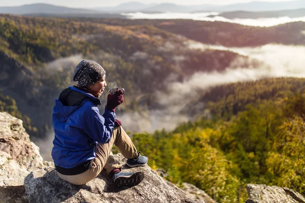 Jovem turista está sentado com uma caneca de chá em um penhasco com vista para as montanhas de outono com nevoeiro. O conceito de um feriado feliz na natureza, Caminhadas — Fotografia de Stock