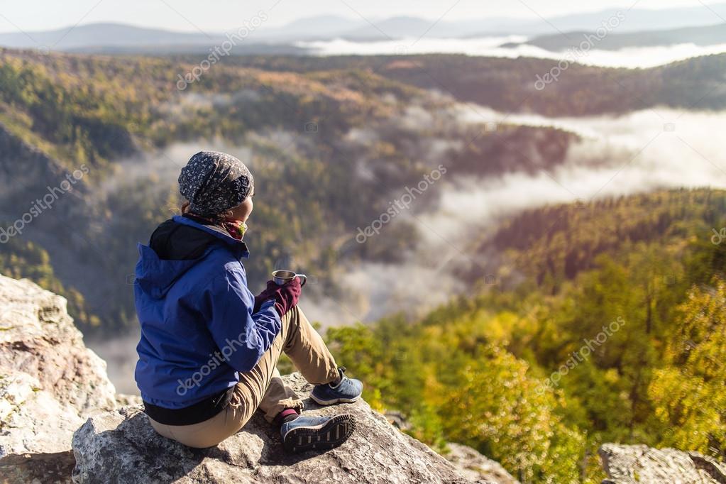 tourist young woman is sitting with a mug of tea on a cliff overlooking the autumn mountains with fog. The concept of a happy holiday in the nature, Hiking