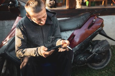 Kırsal kesimde çalışan genç adam motosikletinin yanında oturmuş sigara içiyor ve akıllı telefonuyla oynuyor.