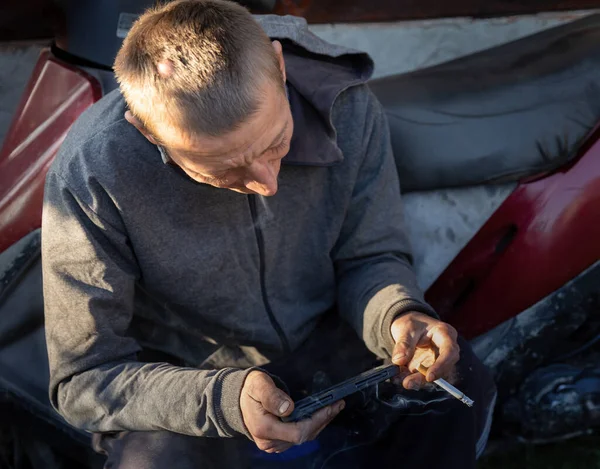 근로자가 오토바이 근처에 앉아서 쉬면서 담배를 피우고 스마트폰을 가지고 — 스톡 사진