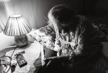 Yaşlı kadın, sehpanın yanındaki karanlık odada, içinde haplar, telefon ve masa lambası olan koltukta oturuyor. Siyah ve beyaz resim