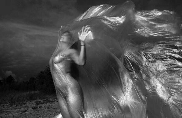 Beautiful body of nude woman on nature background under plastic wrap. Sexy girl behind a polyethylene film by the sea on a windy day.  Fine-art photo.