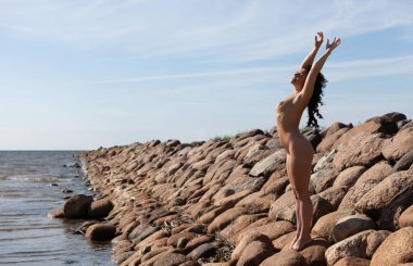 Genç, çıplak bir kadın taştan bir dalganın üzerinde poz veriyor. Çıplak esmer doğanın tadını çıkarıyor.