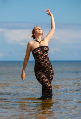 Siyah dantel elbiseli genç, kızıl saçlı kadın sahilde çıplak poz veriyor.