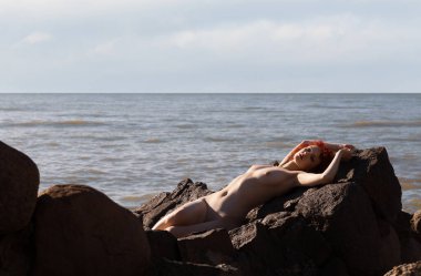 Deniz kıyısındaki eski bir askeri kalenin taşları arasında çıplak kızıl saçlı genç bir kadın.