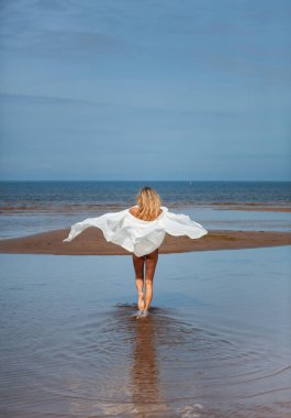 Yaz güneşli bir günde, rüzgârda uçan beyaz bir kumaşla deniz kıyısında poz veren genç, çıplak sarışın. Seksi kadın yaz tatilinden zevk alıyor.