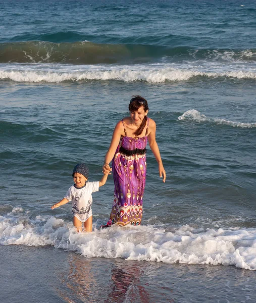 海岸の波の間で彼女の小さな息子と一緒に長い夏のドレスの若い女性が歩く — ストック写真