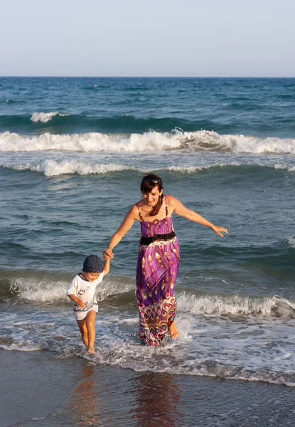 海岸の波の間で彼女の小さな息子と一緒に長い夏のドレスの若い女性が歩く — ストック写真